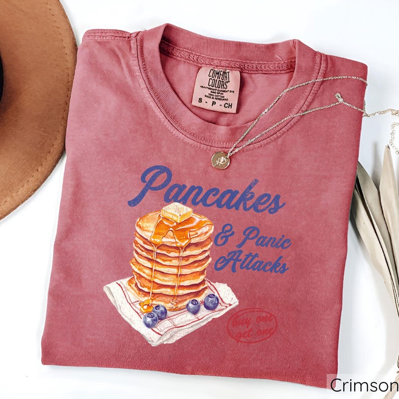 Op de afbeelding: Crimsonrode T-shirt met een afbeelding van een stapel pannenkoeken met siroop en boter, en de tekst "Pancakes & Panic Attacks".