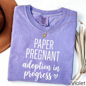 Pode incluir: Uma camiseta Comfort Colors violeta com as palavras "PAPER PREGNANT adoption in progress" impressas em branco. Um colar de ouro com um pingente est&aacute; drapeado sobre a camiseta. Um chap&eacute;u marrom e uma planta seca est&atilde;o no fundo.