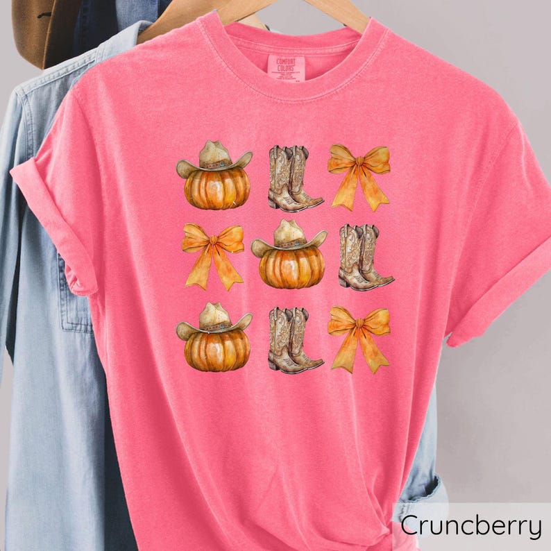 May include: A pink t-shirt with a repeating pattern of watercolor pumpkins wearing cowboy hats, cowboy boots, and orange bows. The shirt has the word "Crunberry" printed on the bottom right.