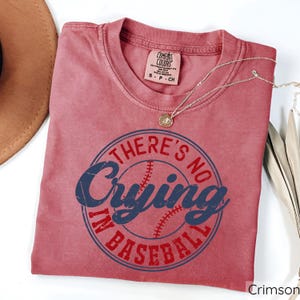 May include: Crimson red t-shirt with a distressed look featuring a white baseball with blue stitching and the text "There's No Crying In Baseball".