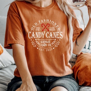 Pu&ograve; includere: Una maglietta arancione con una grafica bianca che recita "Old Fashioned Candy Canes" con illustrazioni di bastoncini di zucchero. La grafica include anche "Kringle Candy Co. Est. 1822".