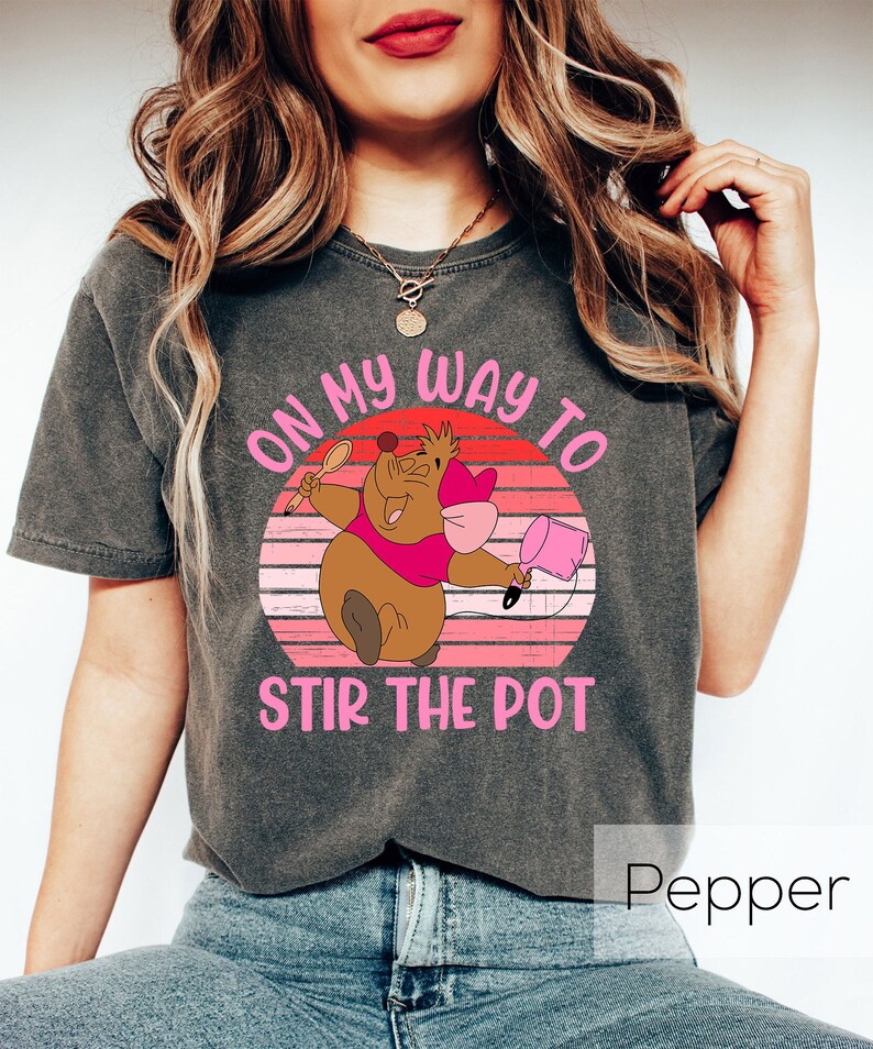 May include: A grey t-shirt featuring a pink and brown cartoon character holding a spoon and a bucket. The text "On my way to stir the pot" is printed on the shirt. The word "Pepper" is printed in white on a black background below the shirt.