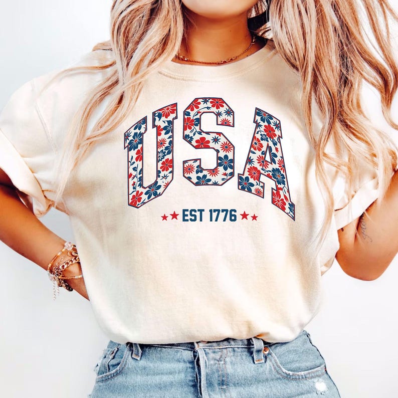 May include: Cream-colored t-shirt with the word "USA" in a floral design of red and blue flowers. Below the word "USA" is the text "EST 1776" with two red stars on each side. The shirt is a crew neck.