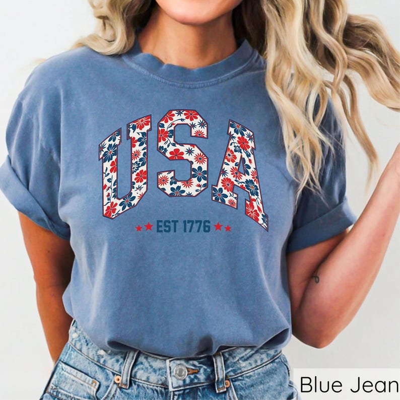 May include: A blue t-shirt with the word "USA" in a floral pattern of red, white, and blue. The text "EST 1776" is below the word "USA" with two red stars on each side. The shirt is made of a soft material.