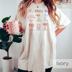 K&ouml;nnte beinhalten: Ein wei&szlig;es Oversize-T-Shirt mit dem Text "It's okay to" und einer Reihe von Illustrationen mit Text darunter. Die Illustrationen zeigen ein Gehirn, eine Sonnenblume, einen Vogel, ein Herz, ein Glas und eine Person. Der Text unter den Illustrationen lautet "need your own space", "make some mistakes", "always be yourself", "not be okay everyday", "not know everything" und "change your mind". Das T-Shirt tr&auml;gt ein Model mit schwarzen Shorts. Der Hintergrund ist eine wei&szlig;e Wand mit einer Pflanze in einem wei&szlig;en Topf.