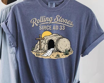 Può includere: T-shirt blu denim con la scritta "Rolling Stones Since AD 33". Il disegno include una croce, una tomba e un sole nascente, suggerendo un tema religioso. La maglietta è appesa a una gruccia di legno.