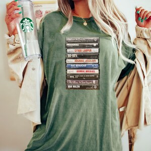 May include: An olive green t-shirt displaying a collection of retro cassette tapes, including artists such as Madonna and Michael Jackson. The shirt has a relaxed fit and a faded look, creating a vintage music aesthetic.