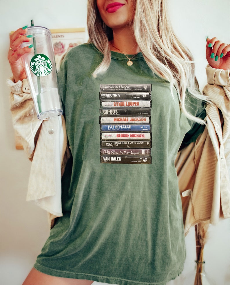 May include: An olive green t-shirt displaying a collection of retro cassette tapes, including artists such as Madonna and Michael Jackson. The shirt has a relaxed fit and a faded look, creating a vintage music aesthetic.