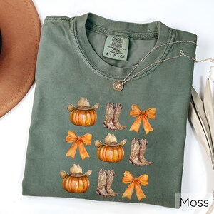 May include: A moss green Comfort Colors t-shirt featuring a design of pumpkins wearing cowboy hats, cowboy boots, and orange bows. The shirt has a crew neck and short sleeves. A silver necklace with a pendant is draped across the shirt.
