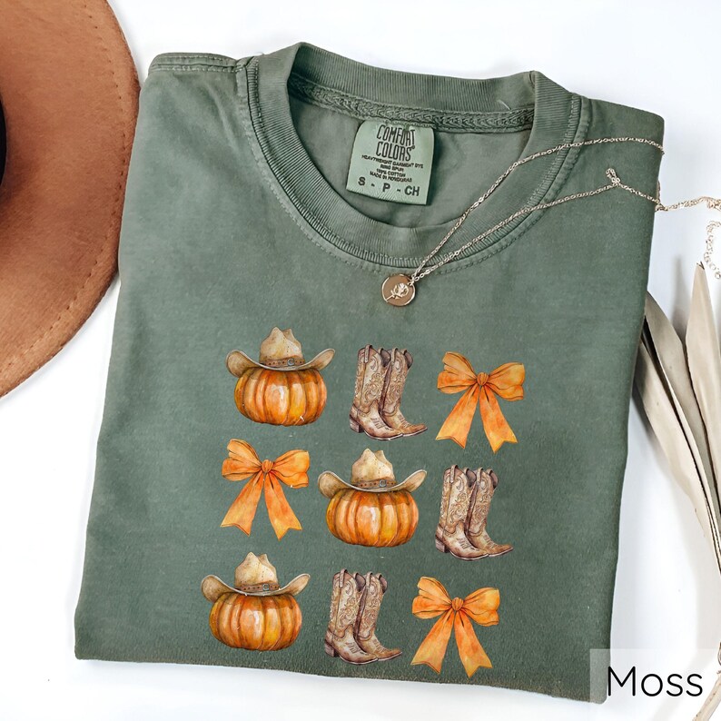 May include: A moss green Comfort Colors t-shirt featuring a design of pumpkins wearing cowboy hats, cowboy boots, and orange bows. The shirt has a crew neck and short sleeves. A silver necklace with a pendant is draped across the shirt.