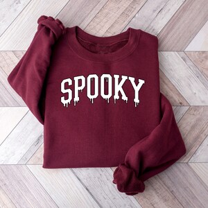 May include: A maroon sweatshirt with the word "SPOOKY" in white, dripping letters. The sweatshirt is folded on a light wood-grain surface. The design has a spooky, Halloween theme.