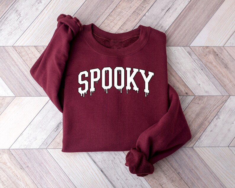 May include: A maroon sweatshirt with the word "SPOOKY" in white, dripping letters. The sweatshirt is folded on a light wood-grain surface. The design has a spooky, Halloween theme.