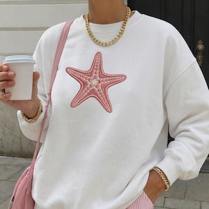 May include: White sweatshirt featuring a detailed pink starfish graphic. The sweatshirt has a crew neck and long sleeves. A gold chain necklace and bracelet are visible. A pink shoulder bag and a white coffee cup are also in the image.