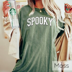 May include: Moss green t-shirt with the word "SPOOKY" in white, dripping font. A clear Starbucks tumbler with a green straw is held in the left hand. The shirt is a relaxed fit, perfect for Halloween or spooky season.