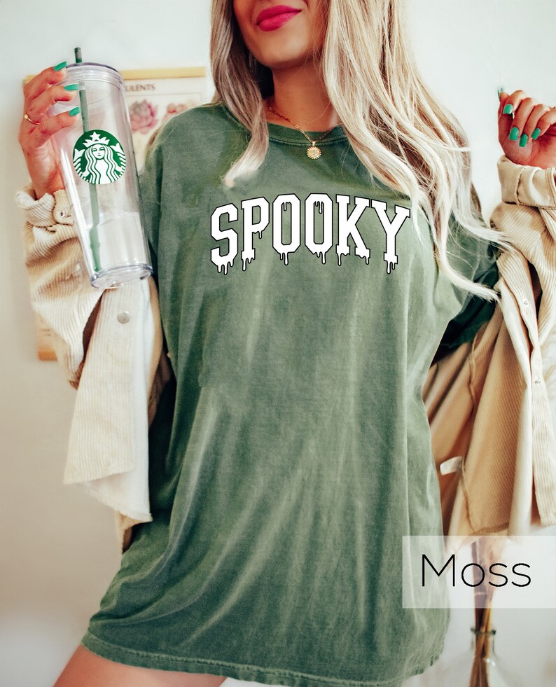 May include: Moss green t-shirt with the word "SPOOKY" in white, dripping font. A clear Starbucks tumbler with a green straw is held in the left hand. The shirt is a relaxed fit, perfect for Halloween or spooky season.