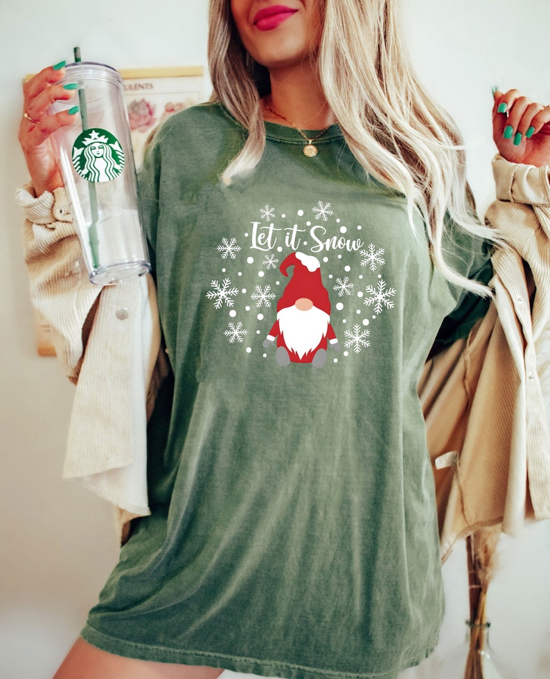 May include: An olive green short-sleeve t-shirt featuring a graphic of a gnome wearing a red hat, surrounded by white snowflakes and the text Let it Snow. The shirt is paired with a clear plastic tumbler with a green straw and a Starbucks logo.
