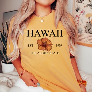 以下が含まれることがあります： 「HAWAII EST 1959 THE ALOHA STATE」という文字と黄色のハイビスカスの花柄がプリントされた黄色のTシャツです。