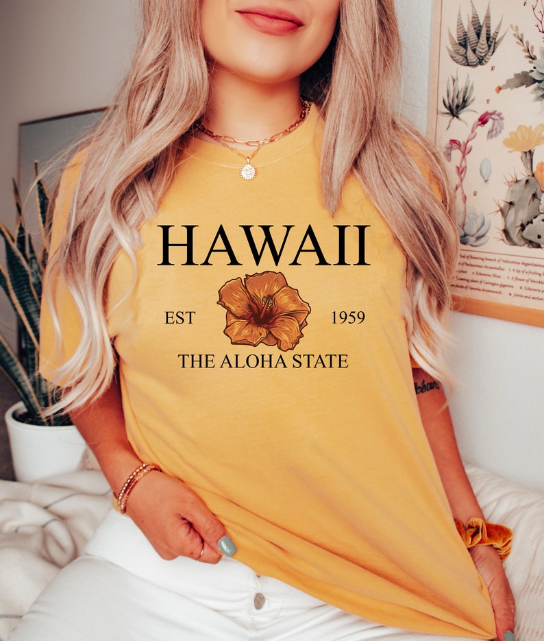 以下が含まれることがあります： 「HAWAII EST 1959 THE ALOHA STATE」という文字と黄色のハイビスカスの花柄がプリントされた黄色のTシャツです。