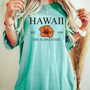 以下が含まれることがあります： ターコイズブルーのTシャツに「HAWAII EST 1959 THE ALOHA STATE」の文字と、茶色のハイビスカスの花柄がプリントされています。