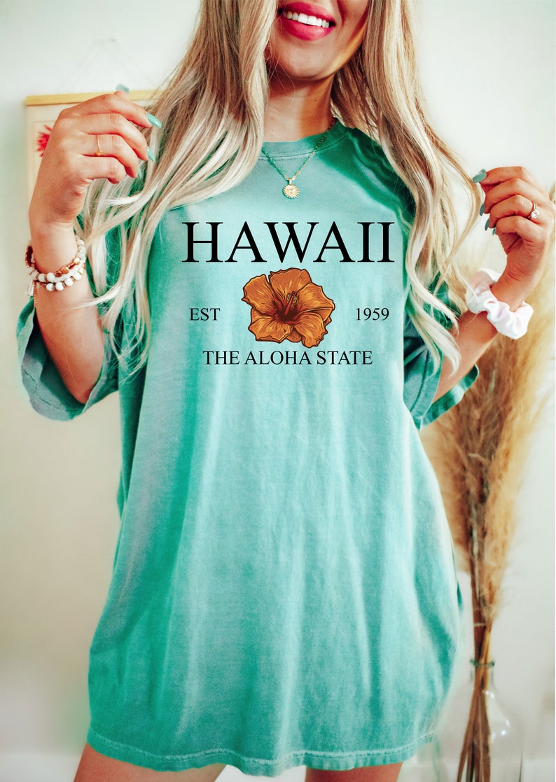 以下が含まれることがあります： ターコイズブルーのTシャツに「HAWAII EST 1959 THE ALOHA STATE」の文字と、茶色のハイビスカスの花柄がプリントされています。