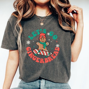 May include: Dark gray t-shirt with the text "LET'S GO GINGERBREAD" in a circular design. The design features a gingerbread man on a skateboard, snowflakes, and a red and green color scheme. The shirt is made of a soft material.
