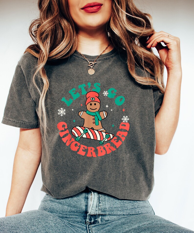 May include: Dark gray t-shirt with the text "LET'S GO GINGERBREAD" in a circular design. The design features a gingerbread man on a skateboard, snowflakes, and a red and green color scheme. The shirt is made of a soft material.