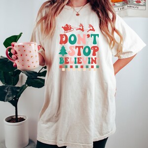 May include: An off-white short-sleeve t-shirt featuring a graphic print with the text "DON'T STOP BELIEVIN'" in red, pink, and green. The design includes a red silhouette of Santa in a sleigh with reindeer, a green Christmas tree, and a checkered border.