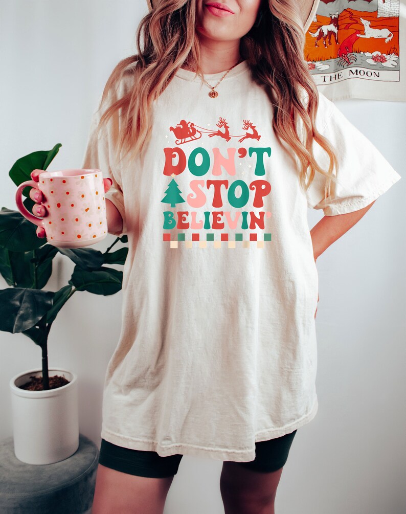 May include: An off-white short-sleeve t-shirt featuring a graphic print with the text "DON'T STOP BELIEVIN'" in red, pink, and green. The design includes a red silhouette of Santa in a sleigh with reindeer, a green Christmas tree, and a checkered border.