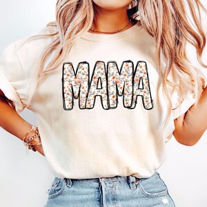 May include: A white t-shirt with a floral print design that says "MAMA" in black outlined letters.