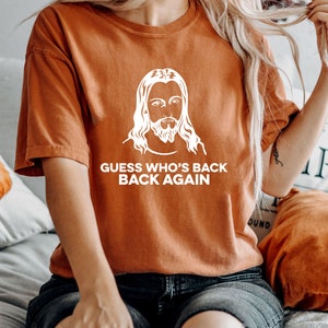 May include: A rust-colored t-shirt with a white graphic of Jesus and the text "GUESS WHO'S BACK BACK AGAIN".