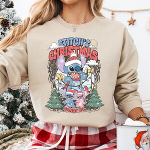 May include: Beige sweatshirt featuring a graphic of Stitch from Lilo & Stitch, wearing a Santa hat and holding Christmas lights. The text "Stitch's Christmas" is above the character, with "Candy Co." below. Two small Christmas trees flank the image.