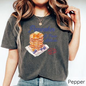 Op de afbeelding: Een donkergrijs T-shirt met een afbeelding van een stapel pannenkoeken met blauwe bessen en siroop. De tekst "Pancakes & Prime Time" staat op het shirt gedrukt.