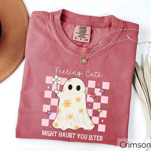 May include: Crimson-colored t-shirt with the text "Feeling Cute Might Haunt You Later." The shirt features a cartoon ghost holding a flower, set against a pink and white checkered background. A gold necklace is draped across the shirt.