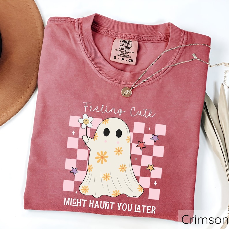 May include: Crimson-colored t-shirt with the text "Feeling Cute Might Haunt You Later." The shirt features a cartoon ghost holding a flower, set against a pink and white checkered background. A gold necklace is draped across the shirt.