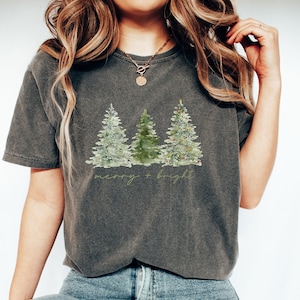 May include: Dark gray t-shirt featuring a watercolor design of three Christmas trees. The trees are in shades of green and silver, with one decorated with ornaments. The text "merry + bright" is written below the trees.