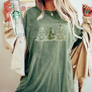 May include: Olive green t-shirt featuring a watercolor design of three Christmas trees. The trees are decorated with snow and ornaments. The shirt has a relaxed fit and a vintage look. The text "merry + bright" is printed below the trees.