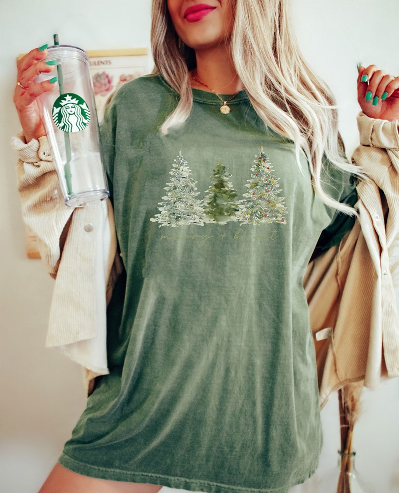 May include: Olive green t-shirt featuring a watercolor design of three Christmas trees. The trees are decorated with snow and ornaments. The shirt has a relaxed fit and a vintage look. The text "merry + bright" is printed below the trees.