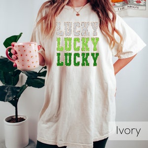 K&ouml;nnte beinhalten: Ein wei&szlig;es T-Shirt mit dem Wort "LUCKY" dreimal in gr&uuml;nem und goldenem Glitzer wiederholt. Das Shirt ist oversized und hat eine lockere Passform. Das Model tr&auml;gt das Shirt mit schwarzen Shorts. Der Text "Elfenbein" ist in der rechten unteren Ecke des Bildes sichtbar.