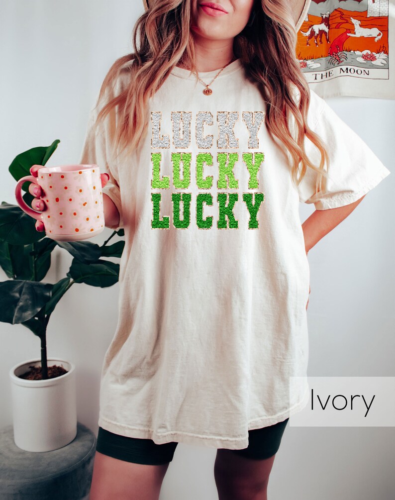 K&ouml;nnte beinhalten: Ein wei&szlig;es T-Shirt mit dem Wort "LUCKY" dreimal in gr&uuml;nem und goldenem Glitzer wiederholt. Das Shirt ist oversized und hat eine lockere Passform. Das Model tr&auml;gt das Shirt mit schwarzen Shorts. Der Text "Elfenbein" ist in der rechten unteren Ecke des Bildes sichtbar.
