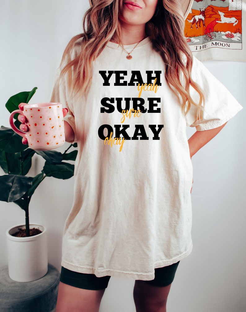 May include: A white t-shirt with black text that reads "YEAH SURE OKAY" with the words "yeah", "sure", and "okay" in a smaller font and a yellow color.