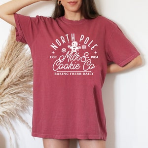 May include: A maroon t-shirt with a white graphic that reads "North Pole Milk & Cookie Co. Baking Fresh Daily" with a gingerbread man and snowflake design. The text also includes "Est 1984". The shirt has short sleeves and a crew neck.