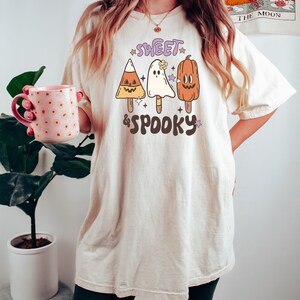 May include: Cream-colored t-shirt with the text "Sweet & Spooky" and illustrations of Halloween-themed popsicles: a candy corn, a ghost, and a pumpkin. The shirt is a relaxed fit.