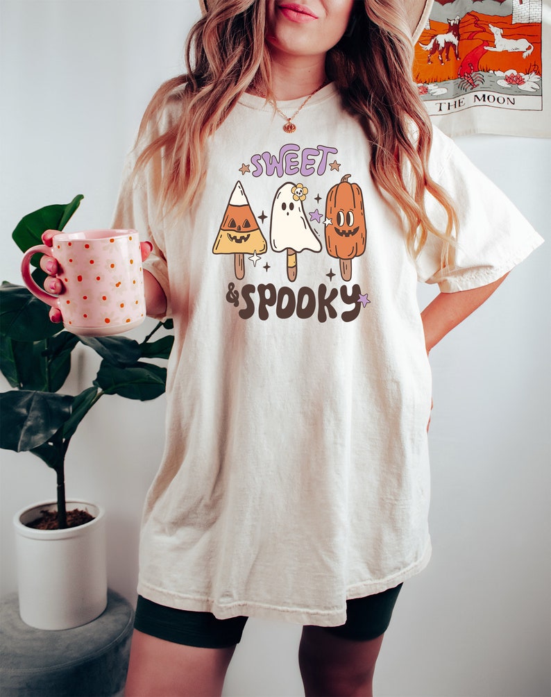 May include: Cream-colored t-shirt with the text "Sweet & Spooky" and illustrations of Halloween-themed popsicles: a candy corn, a ghost, and a pumpkin. The shirt is a relaxed fit.