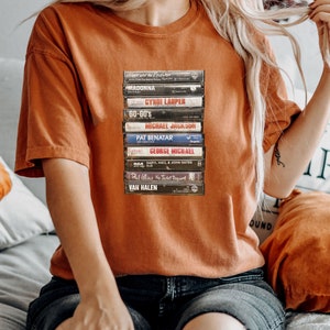 May include: A burnt orange t-shirt with a print of stacked cassette tapes. The tapes feature names such as Madonna, Cyndi Lauper, and Michael Jackson. The shirt has a relaxed fit, ideal for music enthusiasts.