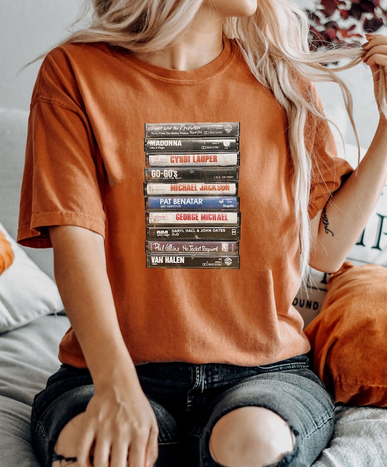 May include: A burnt orange t-shirt with a print of stacked cassette tapes. The tapes feature names such as Madonna, Cyndi Lauper, and Michael Jackson. The shirt has a relaxed fit, ideal for music enthusiasts.