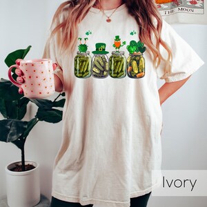 May include: A white t-shirt with a graphic of four pickle jars with leprechauns on top. The jars are filled with pickles and shamrocks. The text "Ivory" is visible in the bottom right corner of the image.