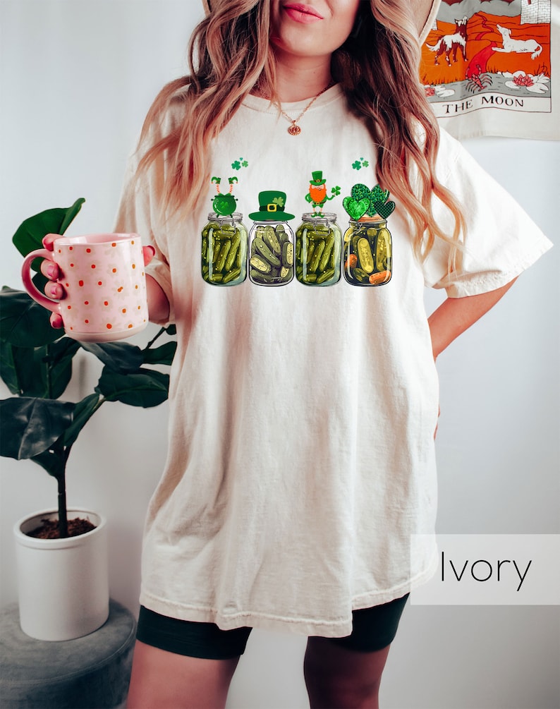 May include: A white t-shirt with a graphic of four pickle jars with leprechauns on top. The jars are filled with pickles and shamrocks. The text "Ivory" is visible in the bottom right corner of the image.