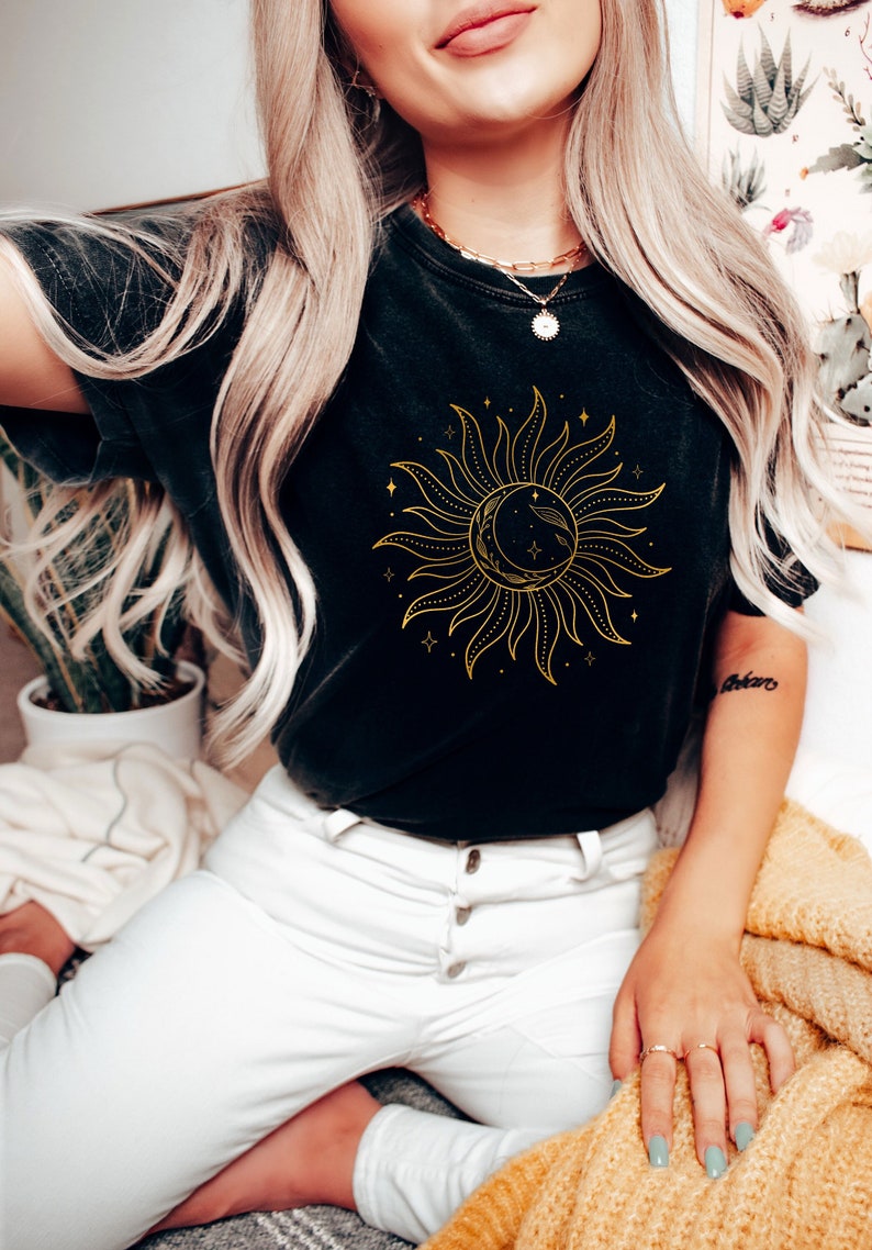 May include: A black t-shirt with a gold sun and moon graphic. The sun has rays and the moon is crescent shaped. The graphic is surrounded by small stars.