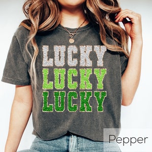 K&ouml;nnte beinhalten: Ein graues T-Shirt mit dem Wort "LUCKY" dreimal in einem gestapelten Design wiederholt. Das Wort "LUCKY" ist in einer glitzernden silbernen Schrift mit gr&uuml;nem Umriss.