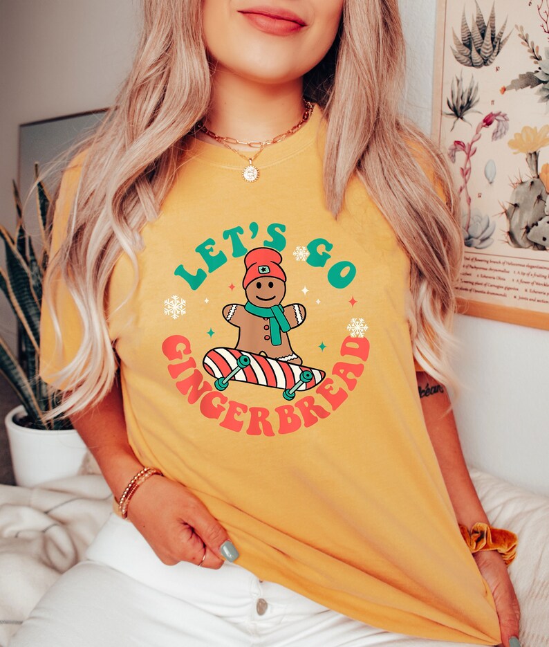 May include: Mustard yellow t-shirt with the text "Let's Go Gingerbread." The graphic features a gingerbread man on a skateboard, surrounded by snowflakes and stars. The gingerbread man wears a red hat and a blue scarf.